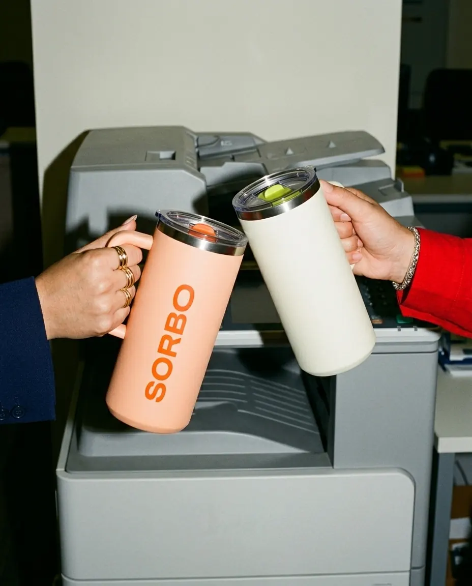 Two hands clink SORBO tumblers in front of a photocopier, under direct flash.